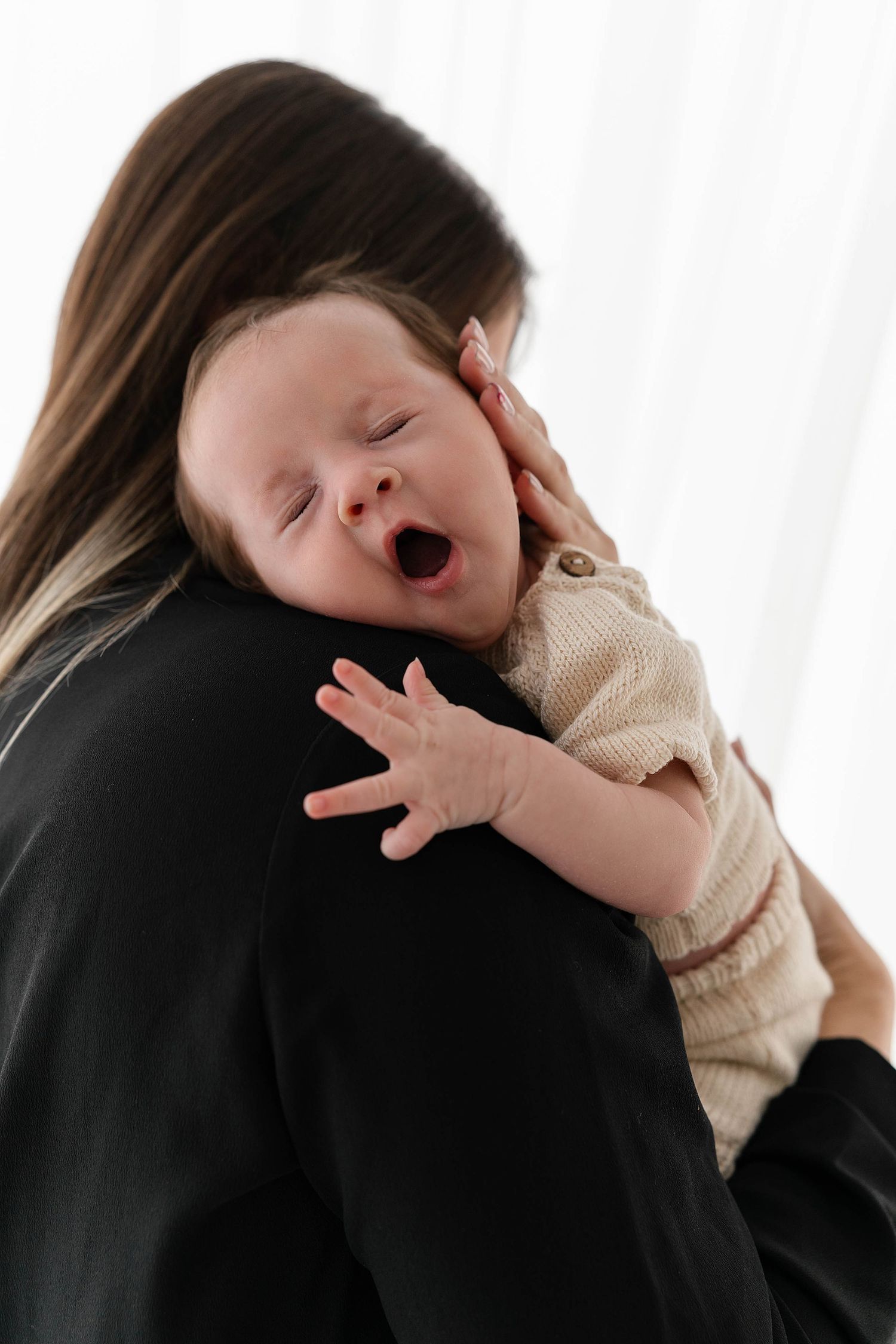 Baby in cream sweater yawns while being held against dark clothing.
