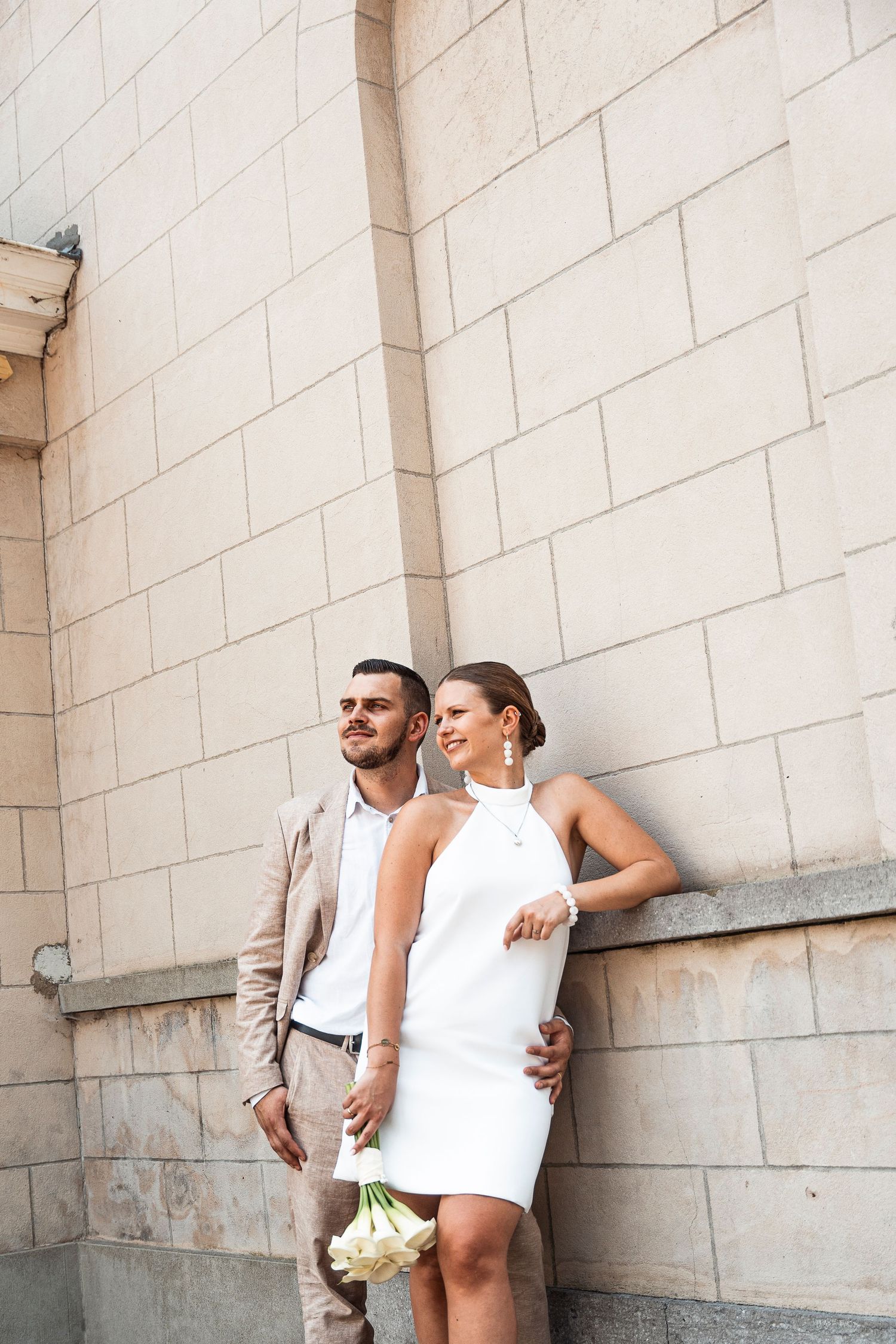 A couple in stylish attire poses against a white stone wall.