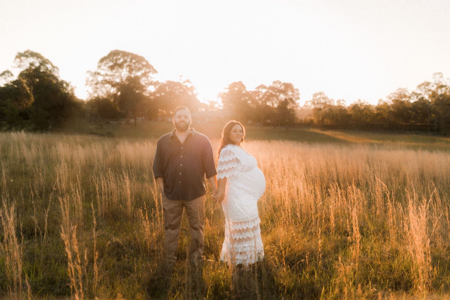 Couple stands together in sunlit field during maternity session.