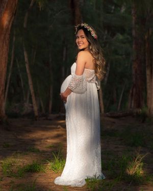 Pregnant woman in white lace off-shoulder gown and floral crown smiling in sunlit forest setting.