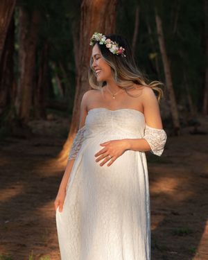 Glowing pregnant woman in white lace dress and flower crown cradling baby bump in woodland setting.