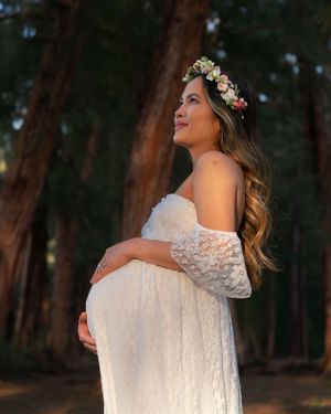 Pregnant woman in white lace maternity gown and floral crown gazing upward among tall forest trees.