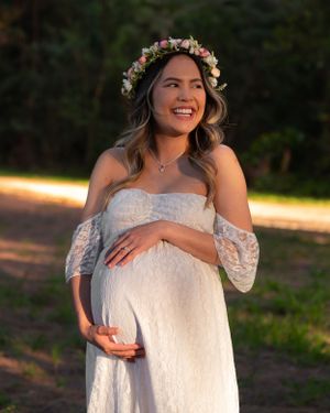 Happy pregnant woman in white lace dress and flower crown laughing while holding baby bump outdoors.