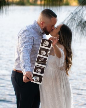 Couple sharing a kiss while holding ultrasound photos during outdoor maternity shoot near a lake.