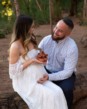 Smiling couple holding tiny brown baby shoes together during outdoor maternity photoshoot on a log.
