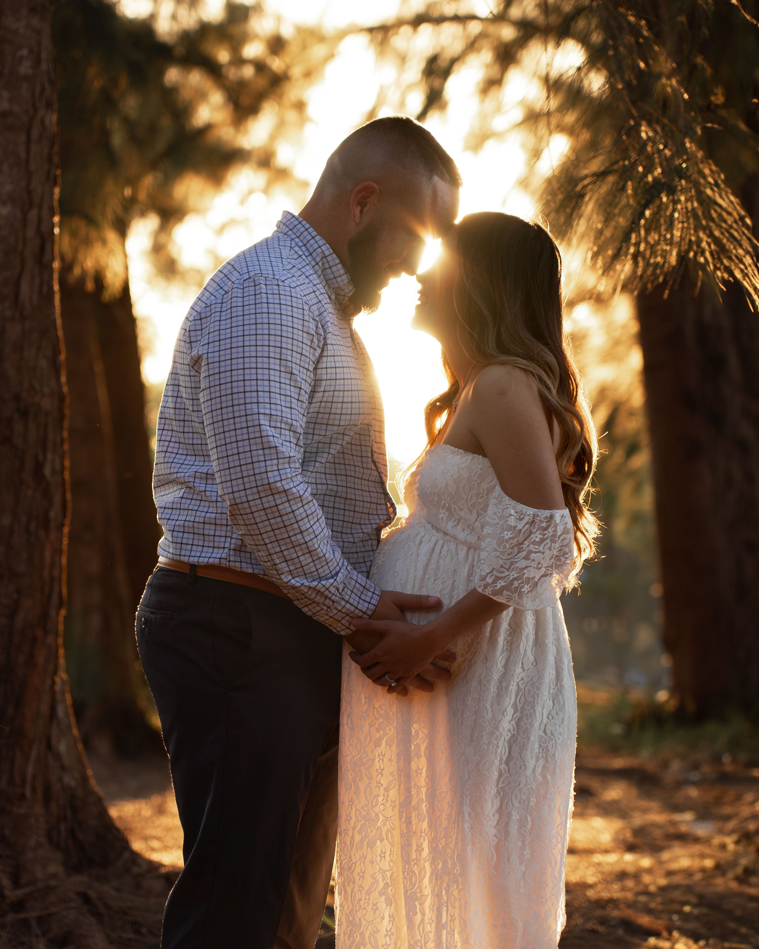 Pregnant couple sharing an intimate moment bathed in warm sunset light among tall trees.