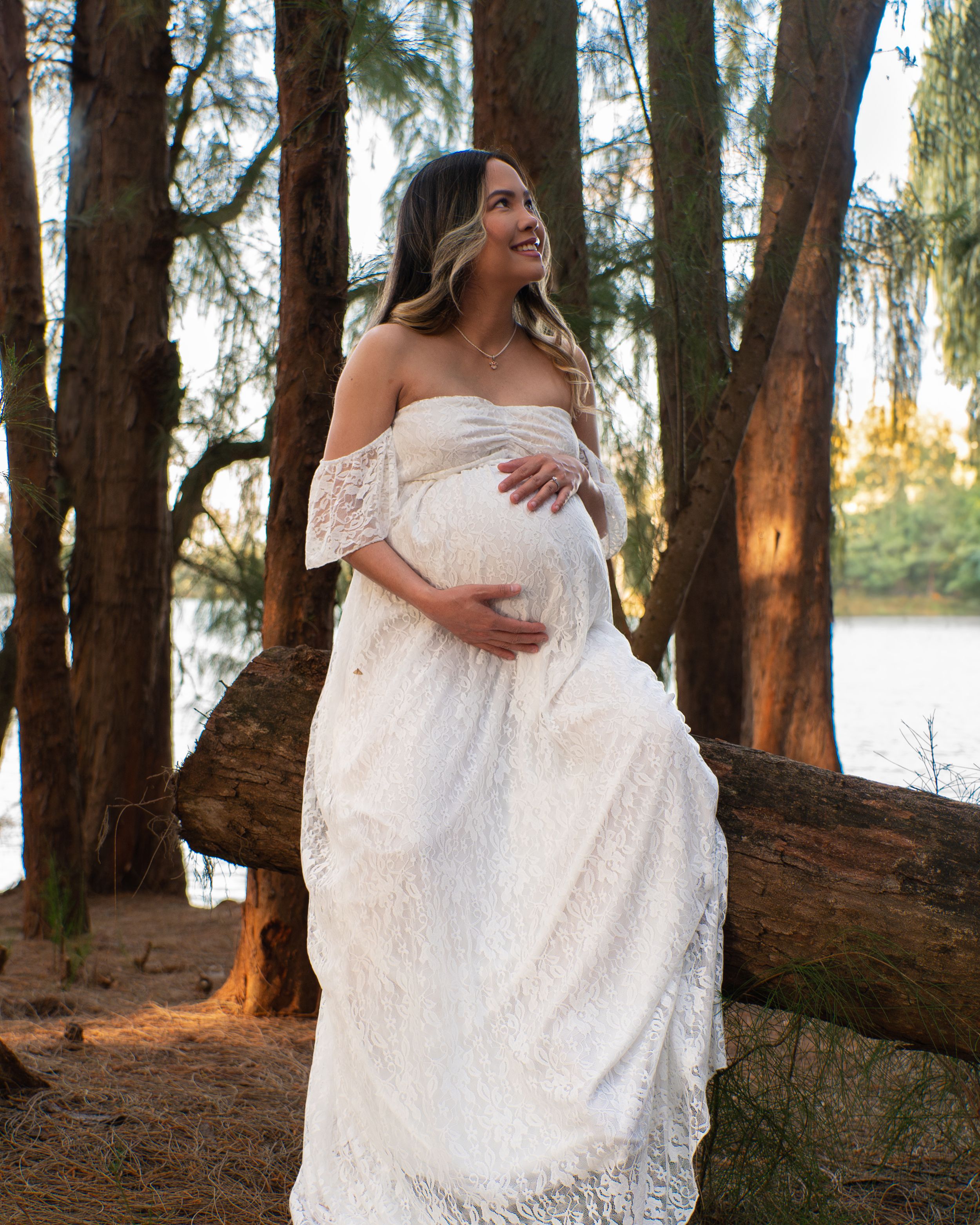 Pregnant woman in white off-shoulder lace maternity gown posing among trees during outdoor photoshoot.