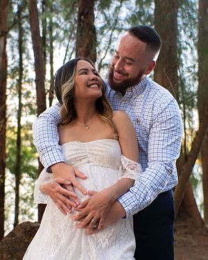 Happy couple embracing during maternity photoshoot in wooded setting, woman in white lace dress.