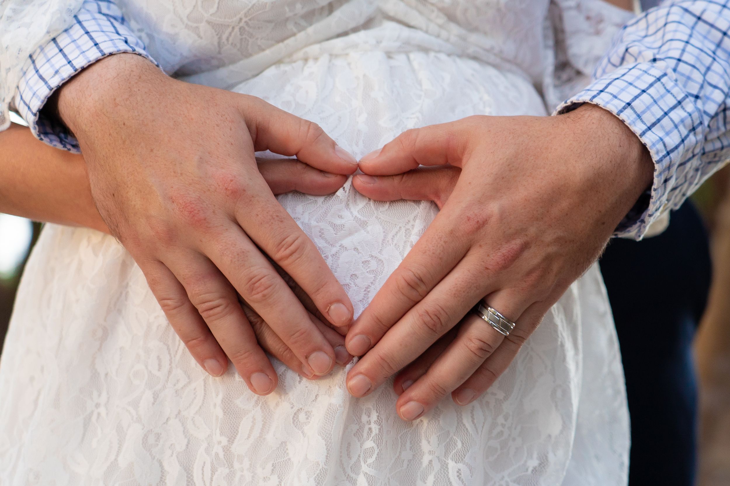 Couple forming heart shape with hands on pregnant belly wearing wedding ring during maternity session.