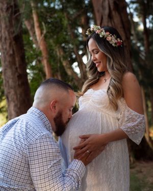 Man kissing pregnant woman's belly as she smiles wearing white lace dress and floral crown outdoors.