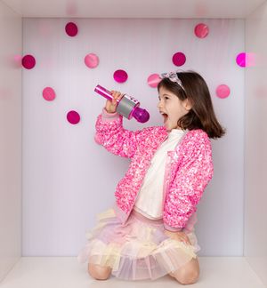 Playful child performs karaoke wearing a pink sequined jacket and tulle skirt against polka dot wall decor.