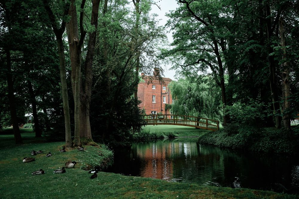 Lush green trees surround a serene pond with a red brick building visible in the distance through the foliage.