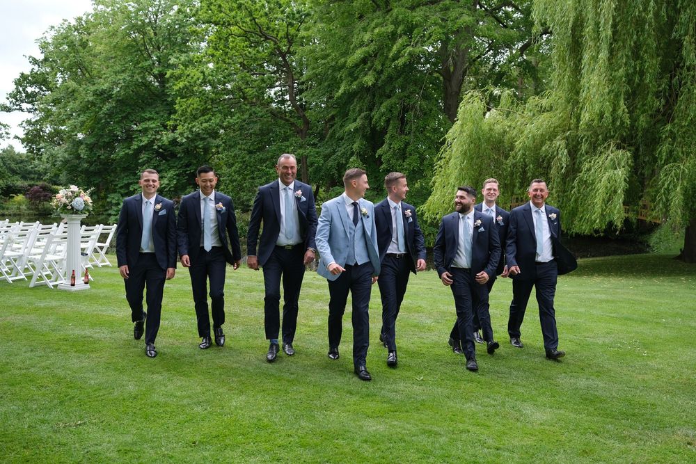 A group of formally dressed groomsmen walk across a lush green lawn with weeping willows and white fencing in background.
