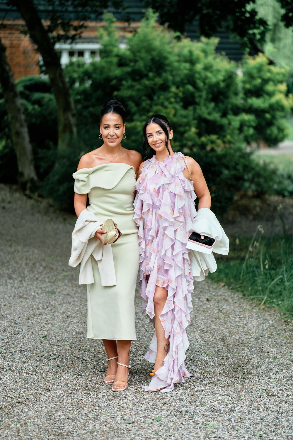 Two guests pose together on a gravel path wearing elegant formal dresses in sage green and lavender with ruffled details.