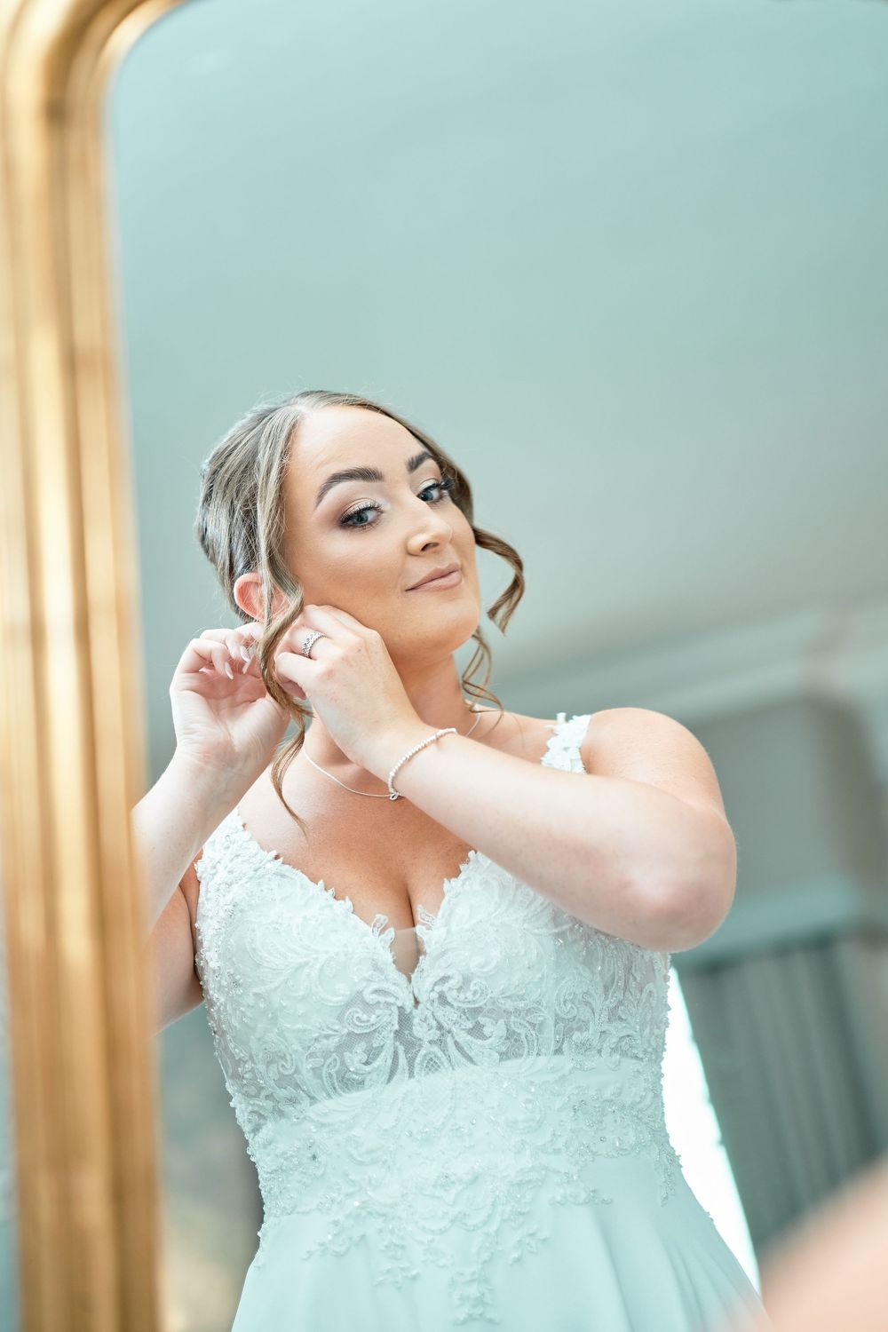 Bride in white lace wedding dress adjusts her earrings while getting ready for the ceremony.