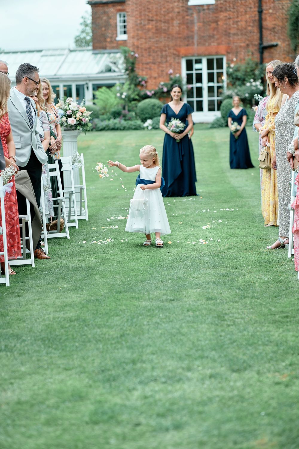 A young child in white walks down a grassy aisle lined with chairs during an outdoor wedding ceremony.