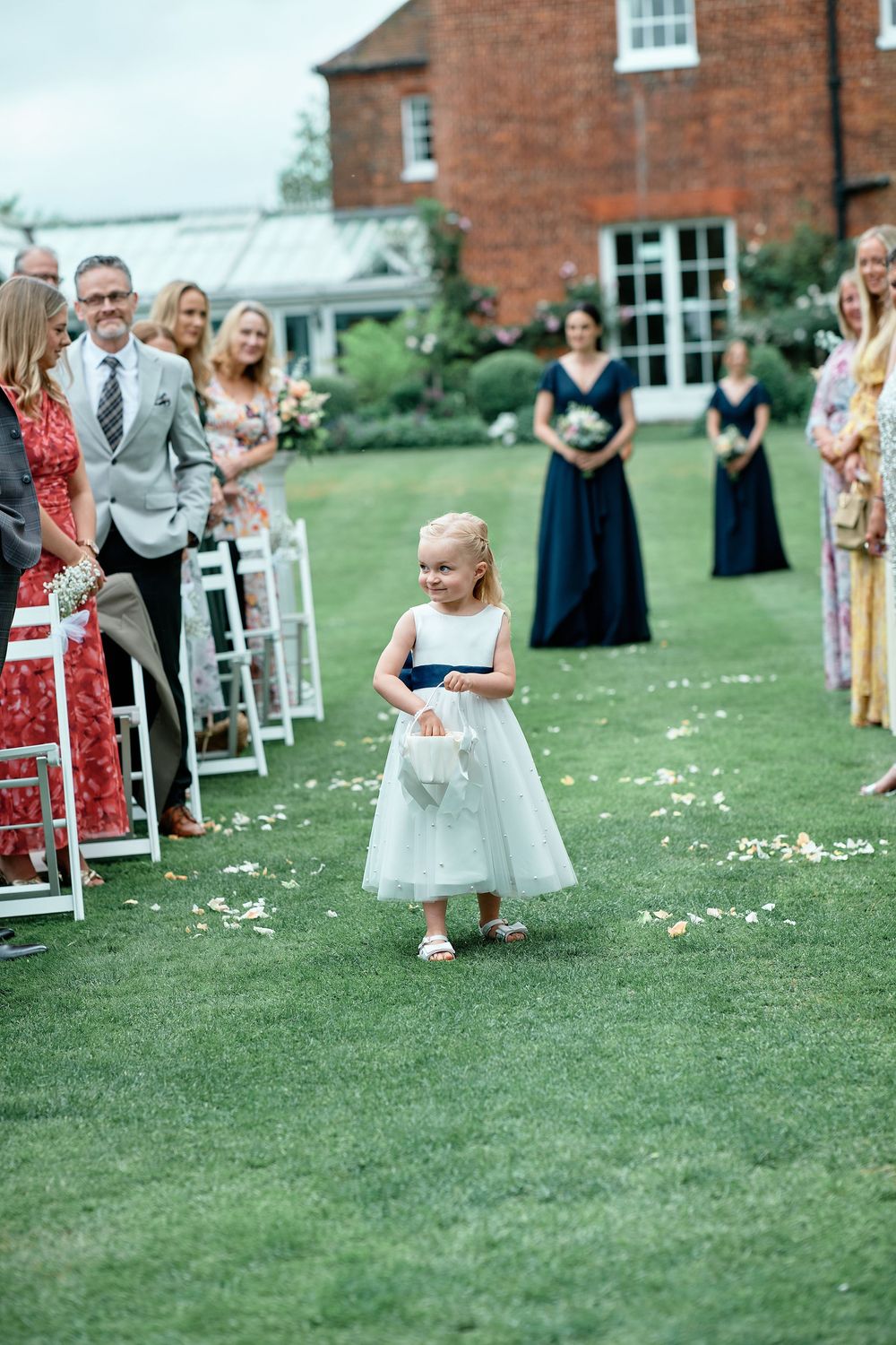 Young flower girl in white dress twirls playfully on grassy lawn at outdoor wedding ceremony while guests watch.