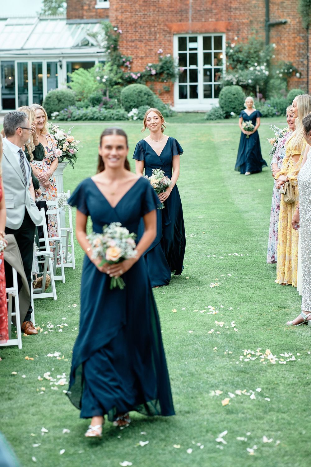 Wedding guests throw flower petals on a green lawn outside a rustic brick building during a celebratory moment.
