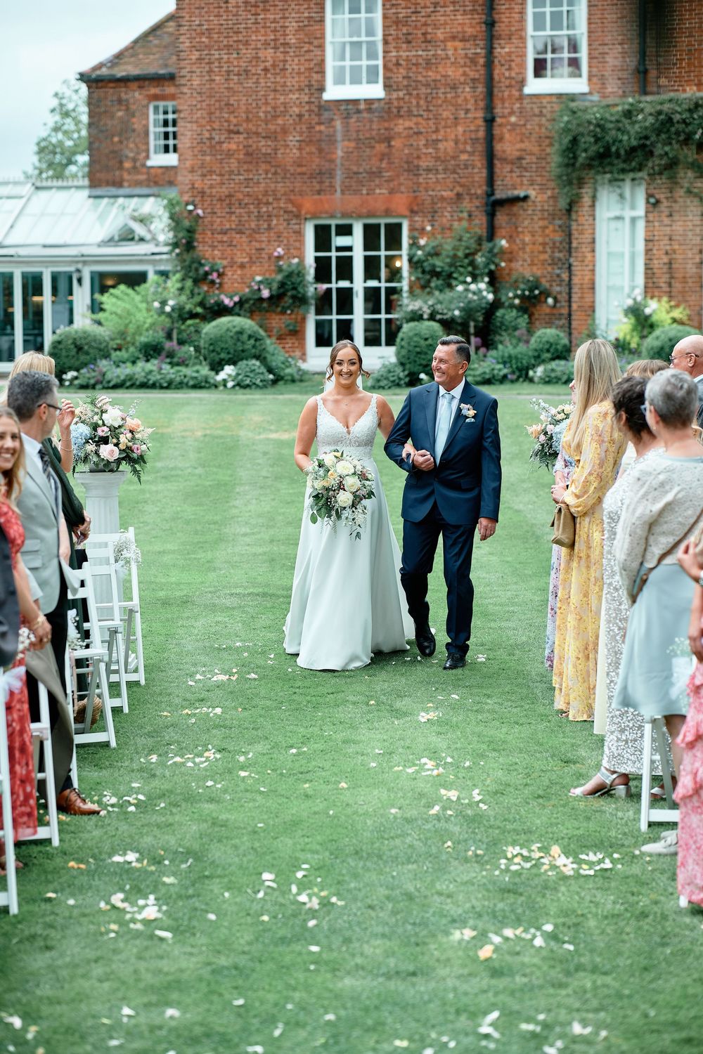 Wedding guests line a garden path during an outdoor ceremony at a classic brick manor house venue.