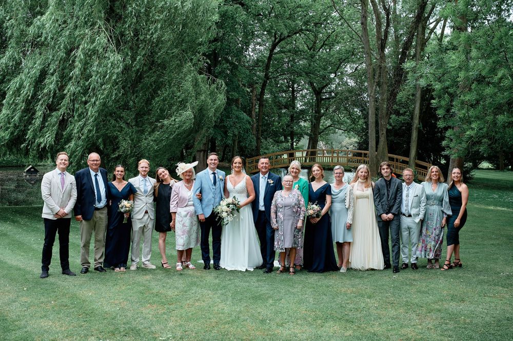 A wedding party group poses together on a lush green lawn surrounded by weeping willow trees and summer foliage.
