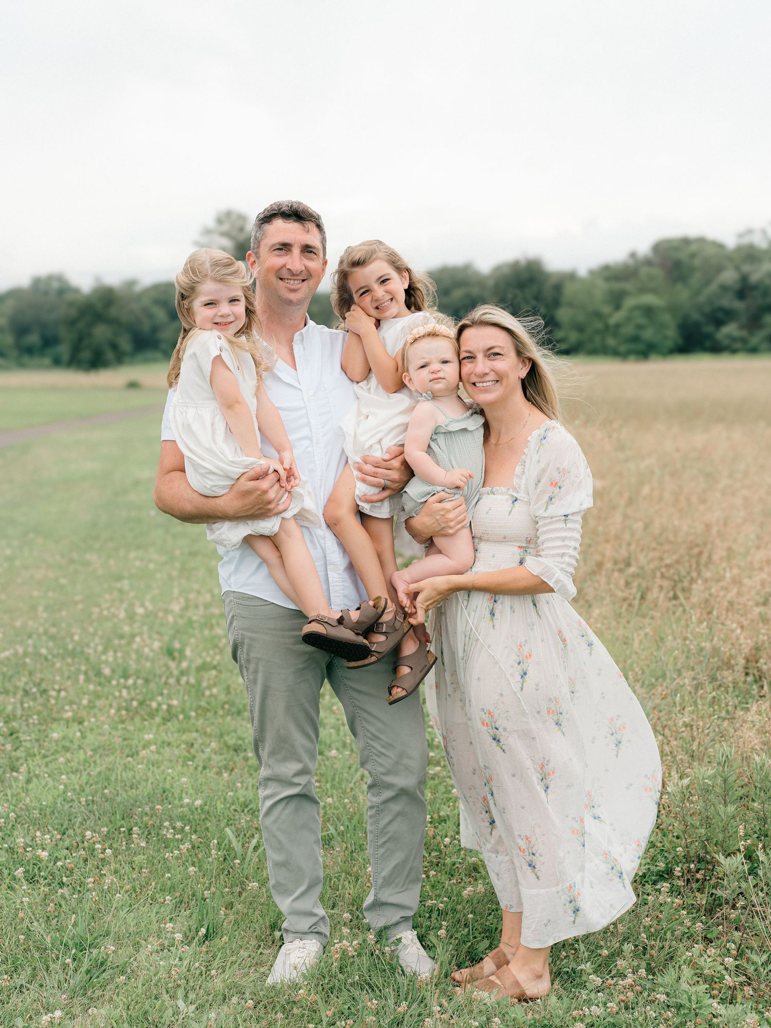 A mother wearing a white floral Doen dress sitting on a stool in a grassy field laughs with her daughters.
