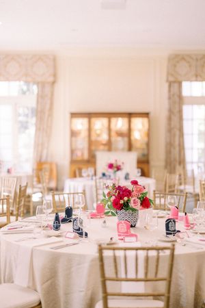 Elegant wedding reception table with pink and red floral centerpiece, white linens, and gold chiavari chairs.