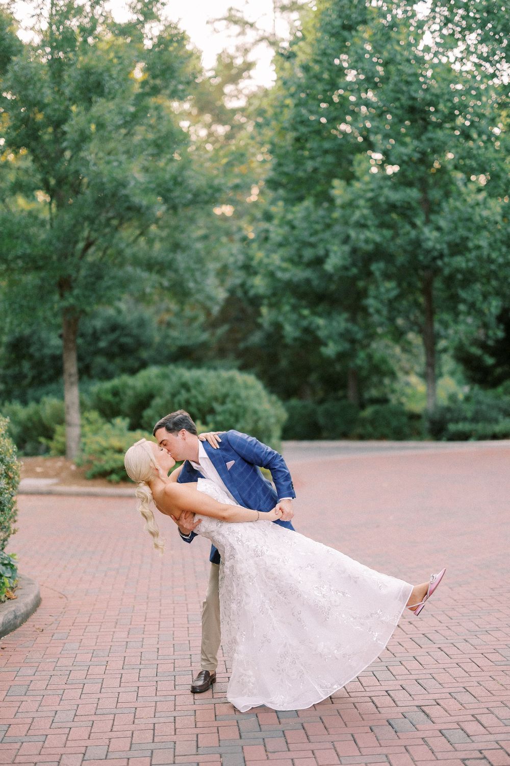 Two people share a romantic dip pose on a brick pathway surrounded by lush green trees at sunset.
