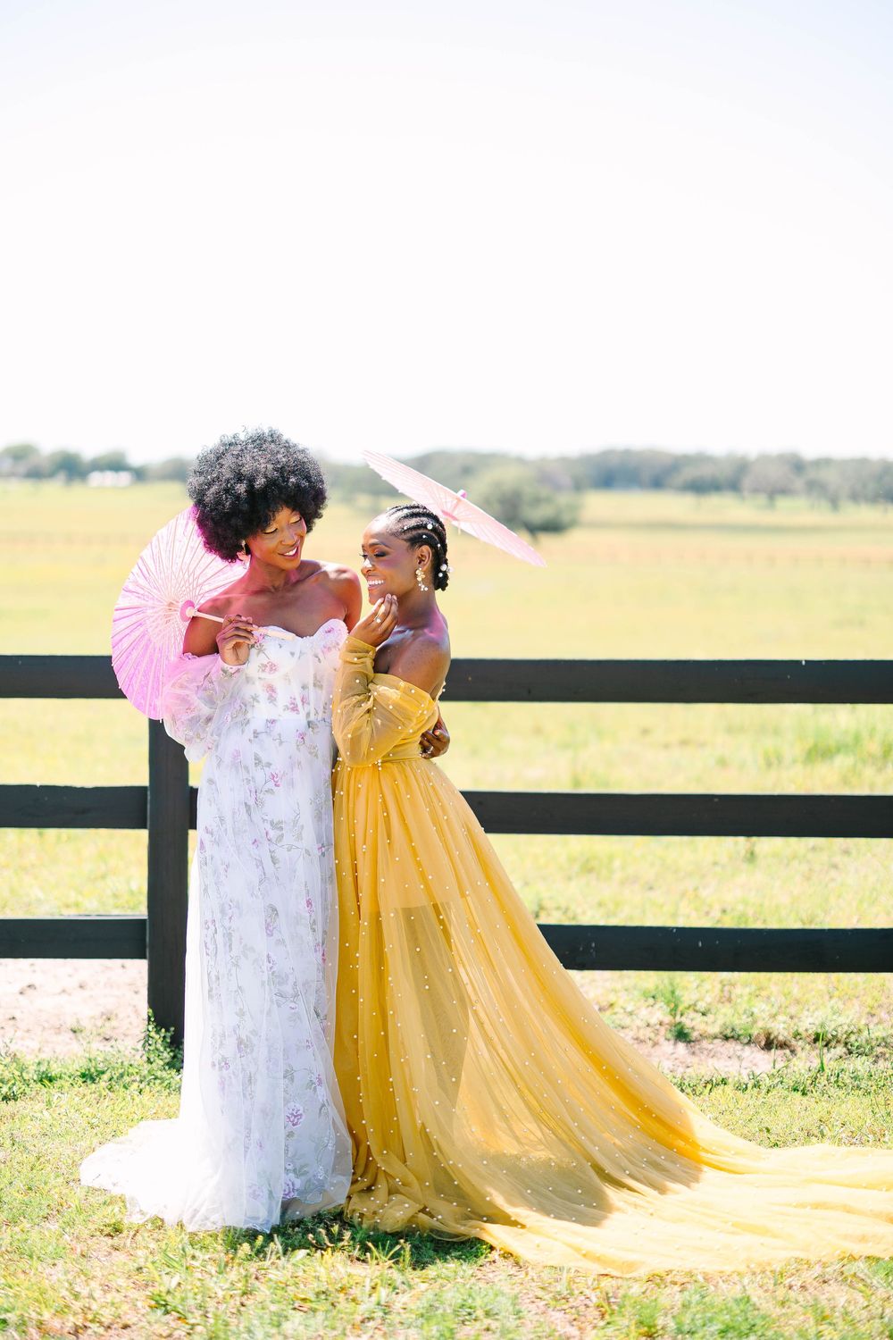 Two people sharing a joyful moment outdoors wearing formal dresses in white and yellow against a wooden fence.