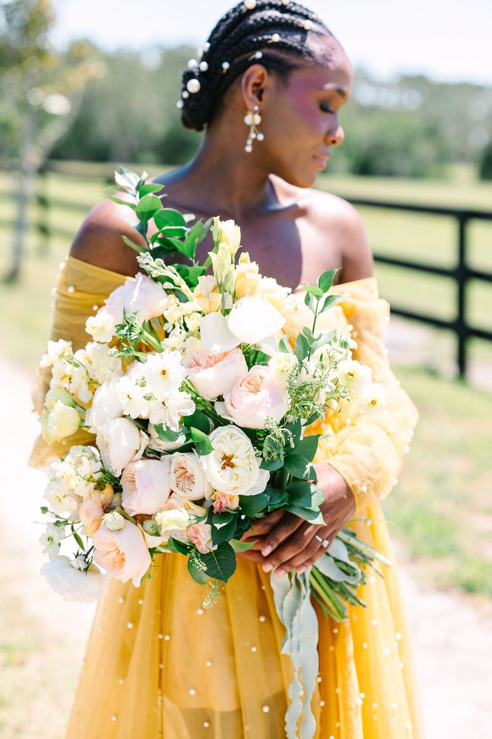 Elegant bridal bouquet of peach and cream flowers paired with a flowing yellow off-shoulder gown in an outdoor setting.