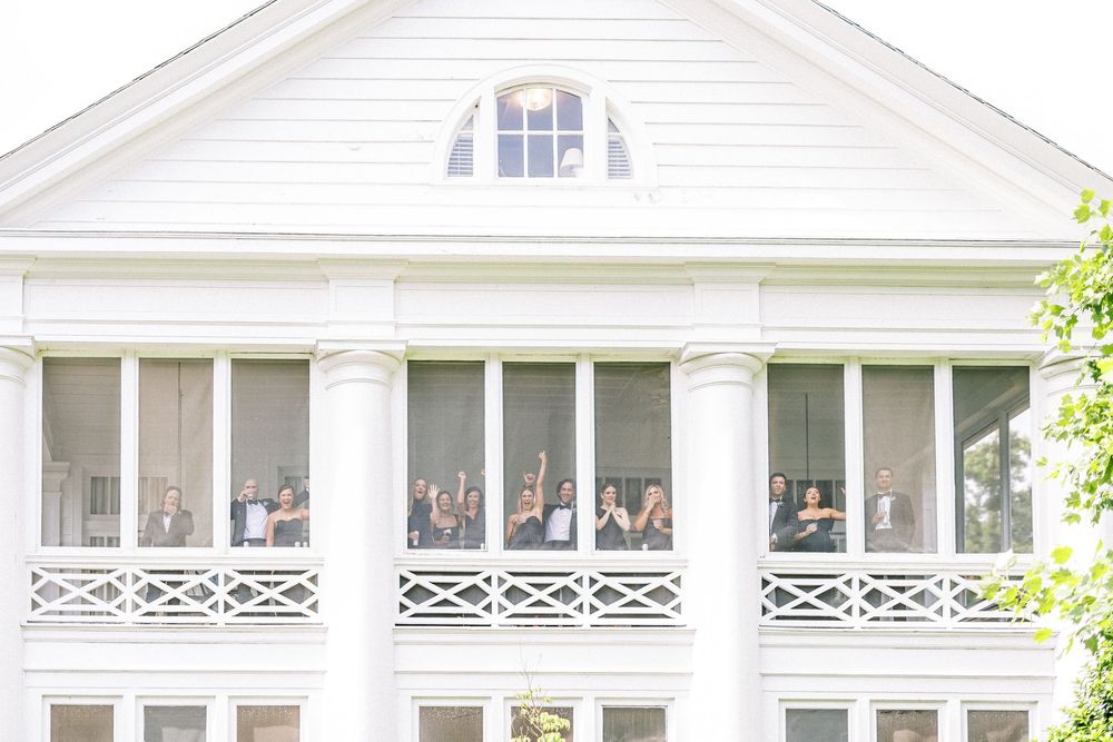 White colonial architecture with covered porch, dormers, and elegant trim details sits among lush greenery.
