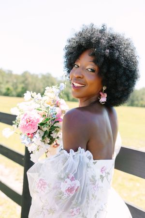 Beautiful bride in off-shoulder floral dress holds pink and white bouquet while smiling on outdoor deck.