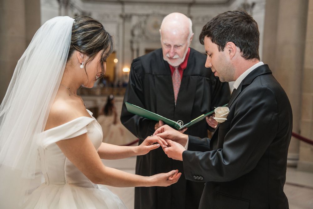 A wedding ceremony takes place indoors as rings are exchanged during an intimate moment.