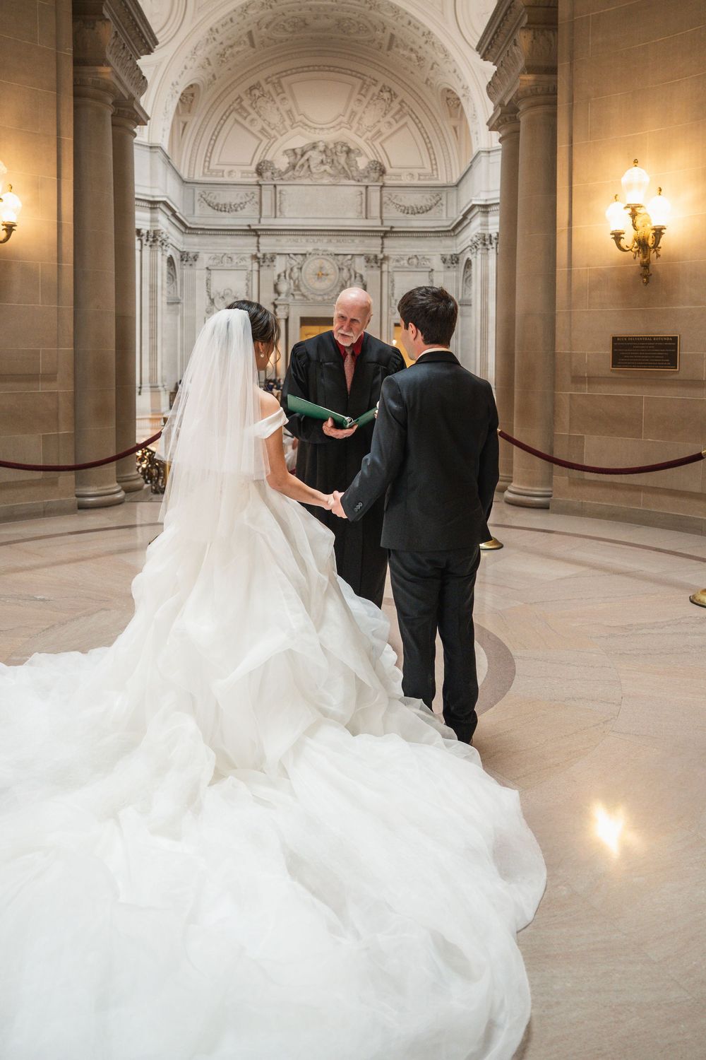 A wedding ceremony taking place inside an ornate classical building with white marble walls and elegant lighting.