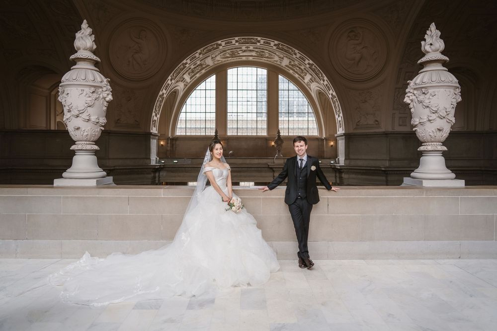 A bridal couple poses in a grand marble-columned hall with ornate architectural details and arched windows.