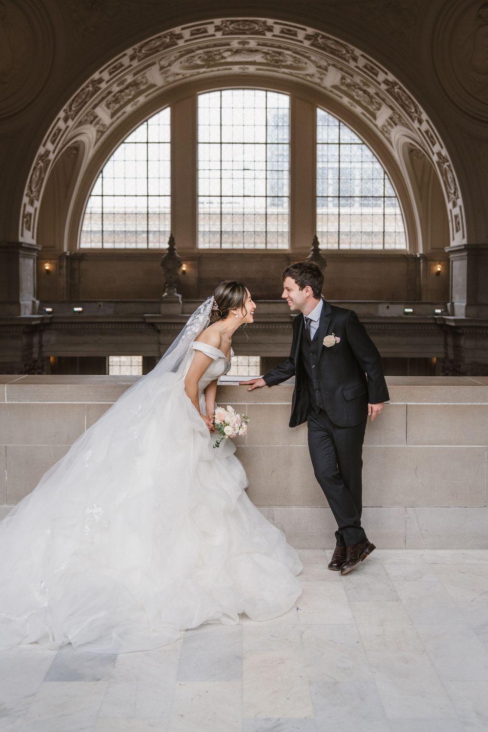 Wedding couple pose on marble steps beneath an ornate arched window in a grand hall.