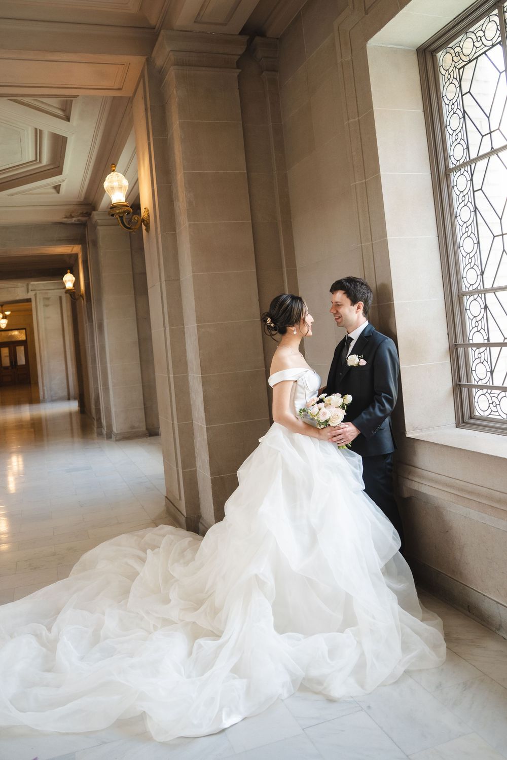 A wedding couple stands in an elegant hallway with marble floors and decorative windows as light streams in.