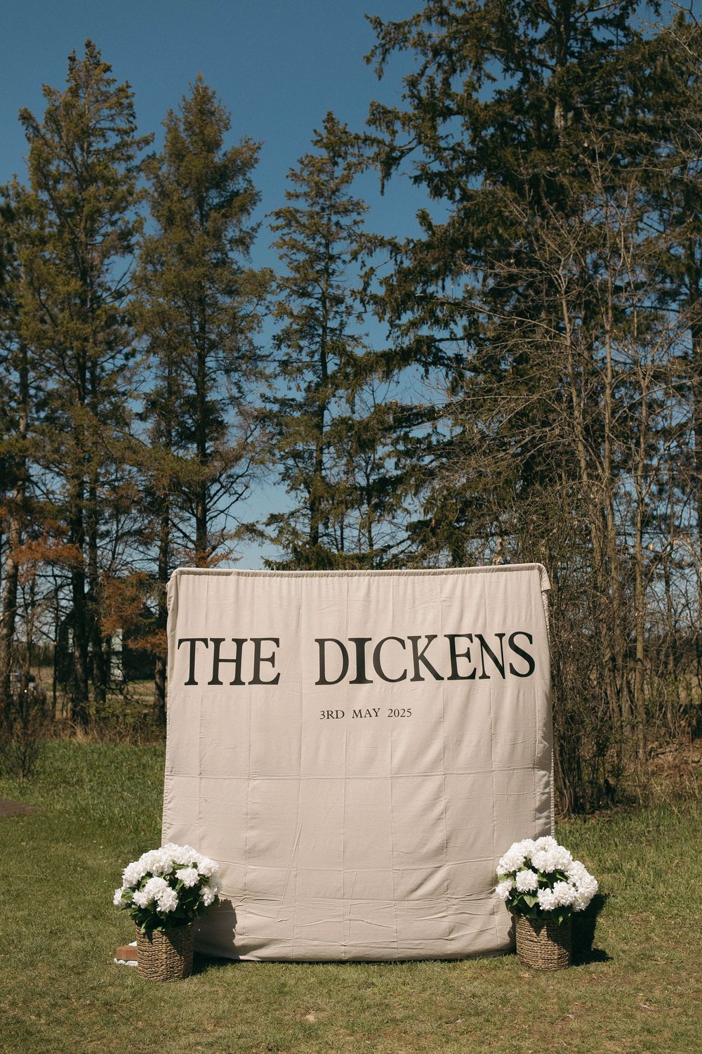 A rustic outdoor wedding sign reading 'THE DICKENS' with white floral arrangements on a grassy field.