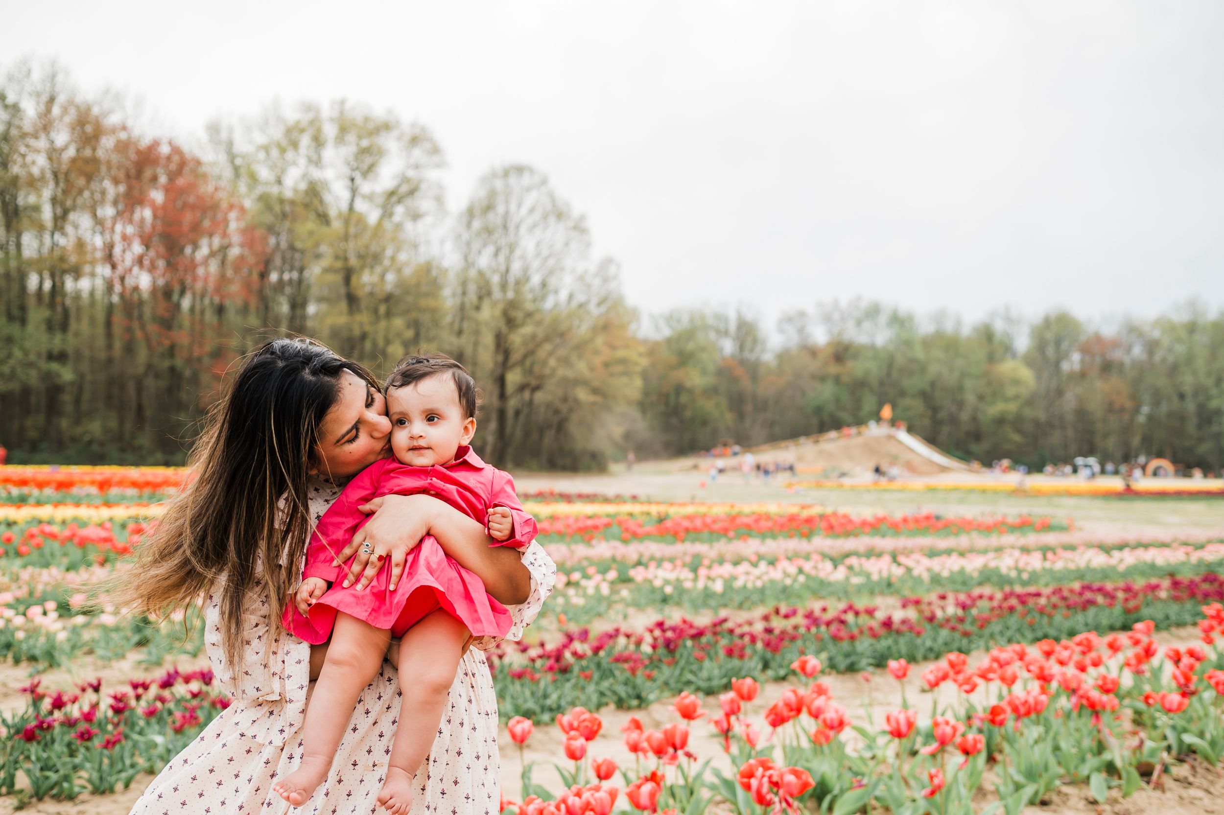 Two people embrace in a floral dress among colorful tulip fields on a spring day with trees in the background.