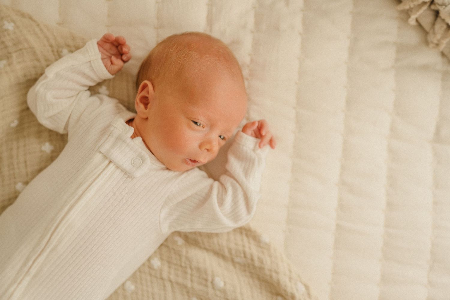 A baby in a white sleeper outfit lies peacefully on cream-colored bedding.