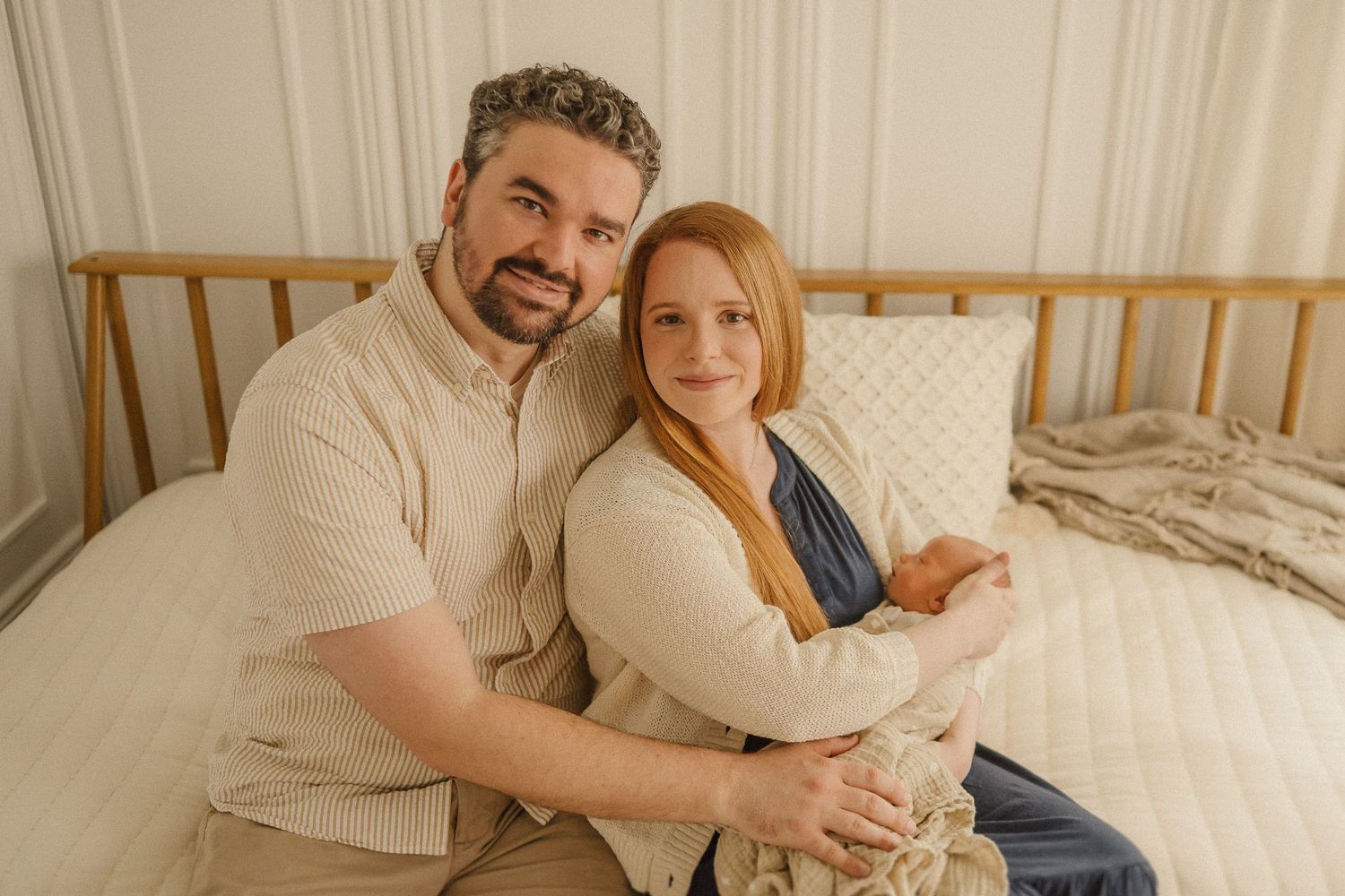 A couple in neutral-toned clothing sits together on a bed against a light backdrop.