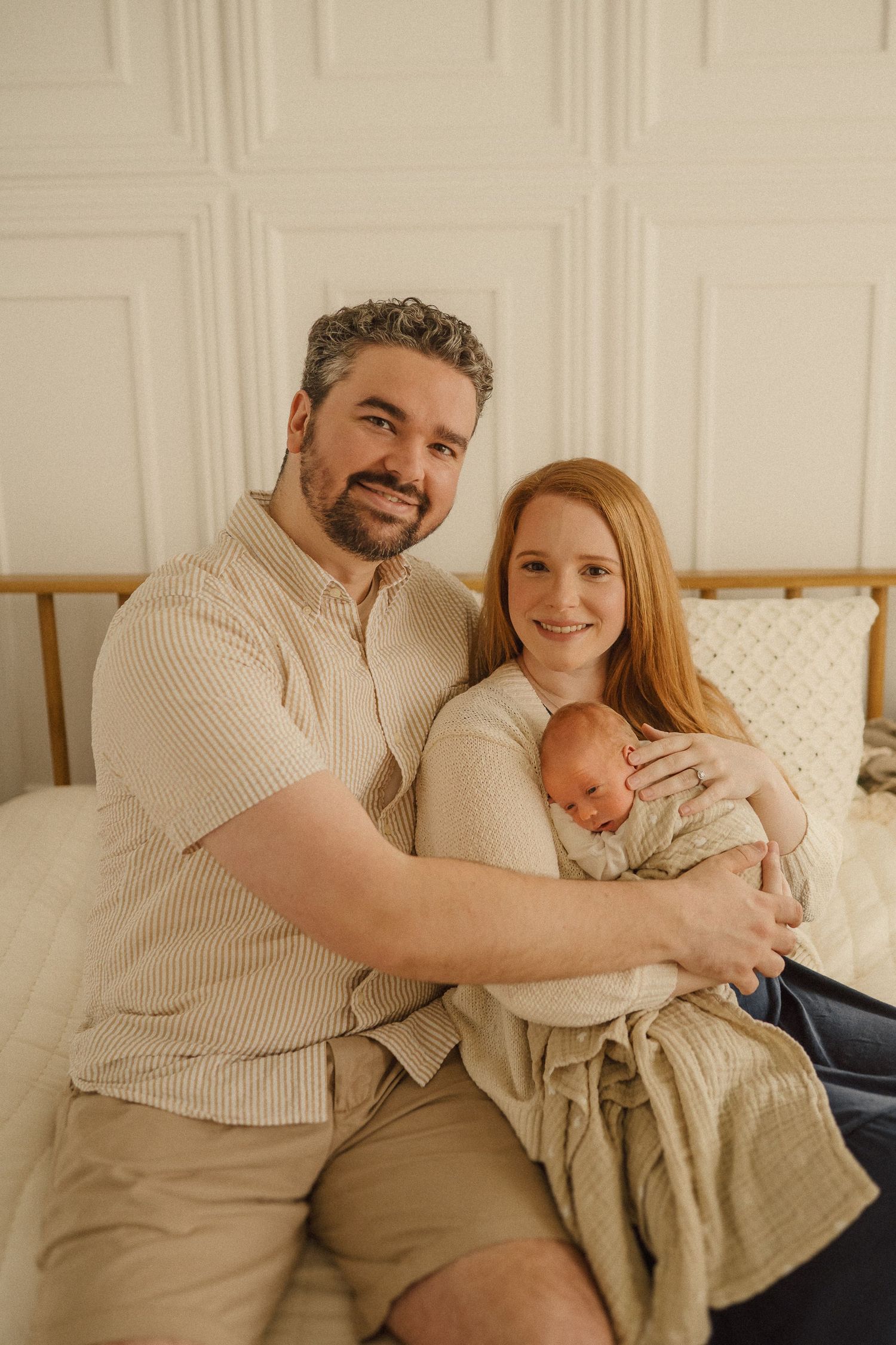 Family photo of parents sitting on bed holding their newborn baby in a cozy neutral-toned setting.