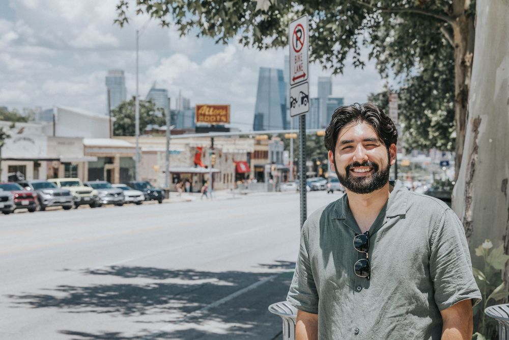 Casual person in a grey henley shirt smiles while standing on a sunny city street.