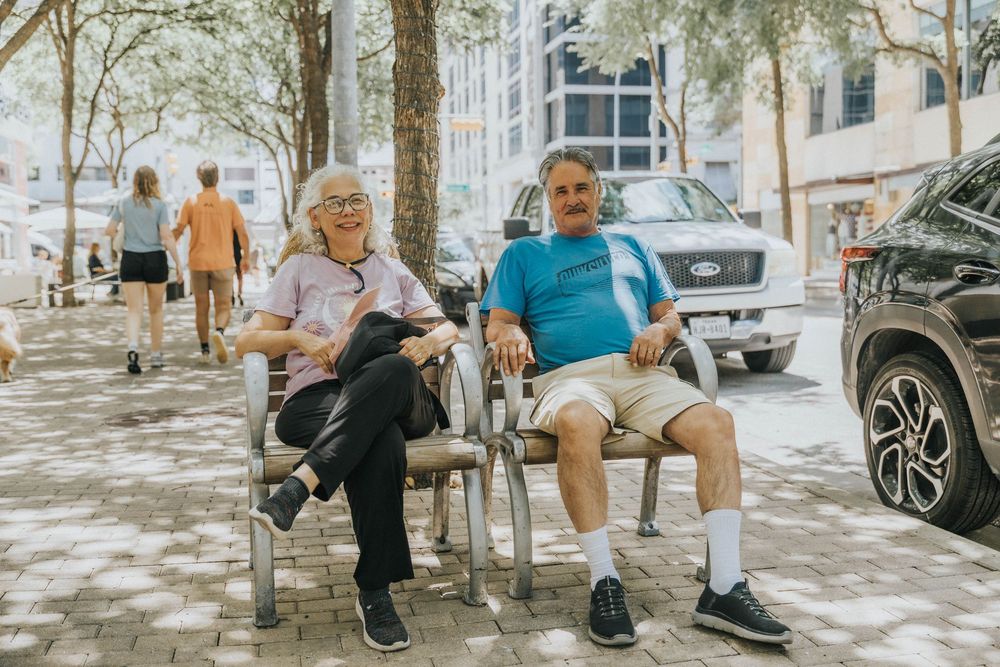 Two people relax on chairs along a tree-lined urban sidewalk on a sunny afternoon.