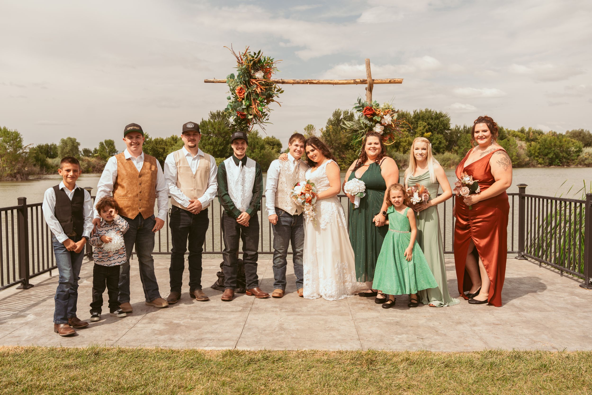 Wedding party stands lined up outdoors under a wooden arch decorated with colorful flowers and greenery.