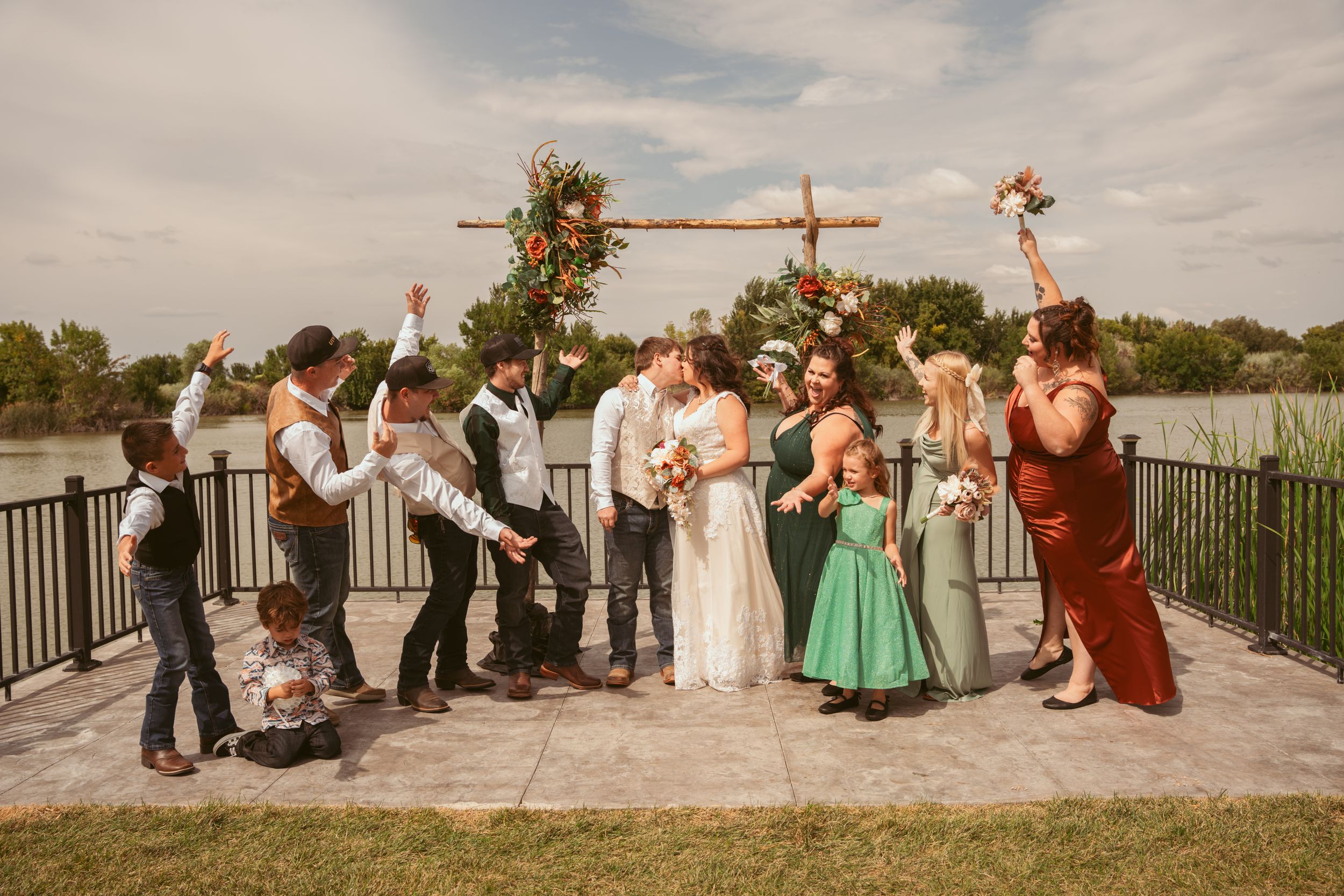 Wedding party gathered under rustic wooden arch with floral decorations by lakeside railing.