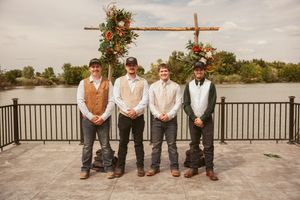 Four groomsmen in coordinated attire stand in a row under rustic wooden arch with water view background.