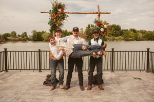 Three groomsmen pose playfully under a rustic floral arch on a lakeside deck.