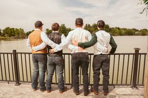 Groomsmen in vests and jeans looking out over lake while leaning against railing.