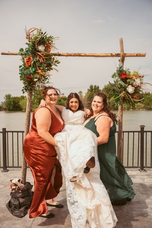Wedding party posing under rustic floral arch against lake backdrop.