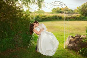 A romantic wedding moment captured under a decorative garden arch in a sunlit field on a bright summer day.